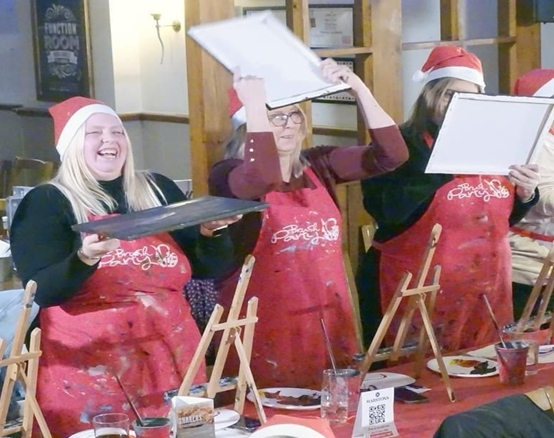 Three people in red aprons laughing behind easels while painting in a pub